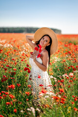 Portrait of a beautiful girl in a field with poppies and daisies . She's wearing a big straw hat...