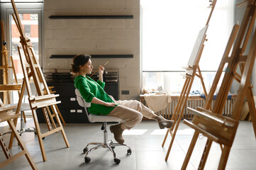 Female artist with paintbrush in hand looking seriously on her own picture