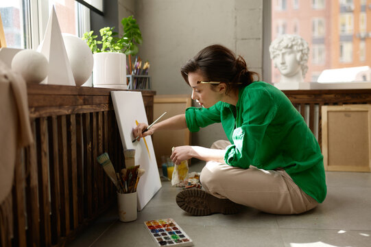 Concentrated woman artist drawing picture sitting on floor in her own art studio