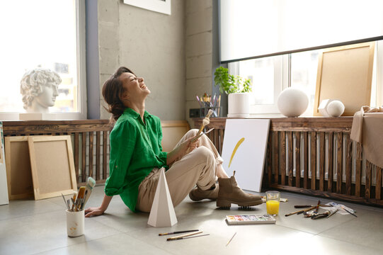 Young female artist laughing loudly sitting on floor at home art studio