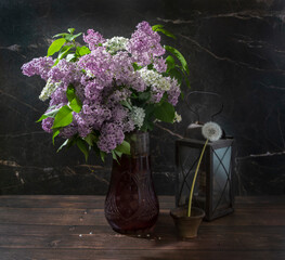 Still life with lilacs in a glass vase with a white dandelion and an antique lamp on a dark marble background. The concept of a moment of life.