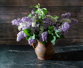 Still life with lilacs and white bird cherry in an old clay vase on a dark marble table. Concept aroma of life.