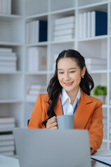 Asian businesswoman sits in a office working on laptop and enjoys a coffee.