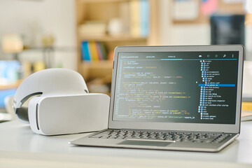 Close-up of laptop with codes on the screen and VR glasses on the table in IT office