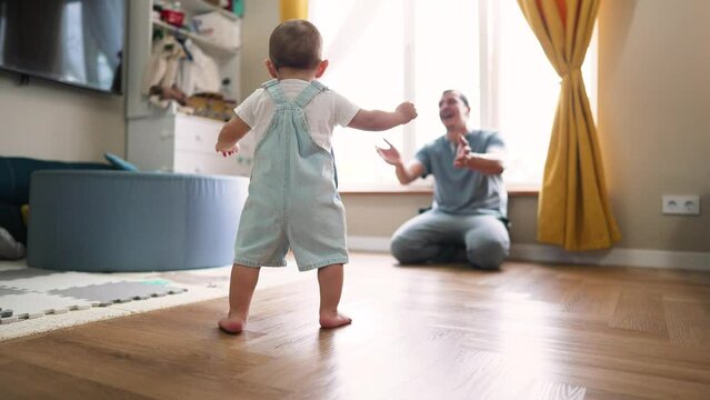 Baby's first steps. Father helps encourages Toddler to take bold first steps at home. Parent lends helping hand for teaches of baby. Toddler walk at home. Baby first active steps on father's day