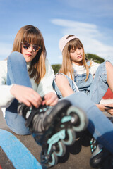 Close up of two teenage blonde sisters sitting adjusting their inline skates in a park. Healthy...