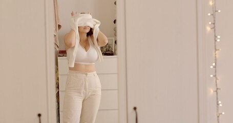 young woman in white puts sweater in front mirror home. Wearing a well-fitting bra during the day might be helpful give extra support so breasts don become sore.  - obrazy, fototapety, plakaty