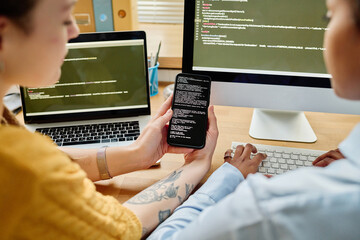 Rear view of colleagues writing security codes in team, they sitting at table with computers and examining code on smartphone
