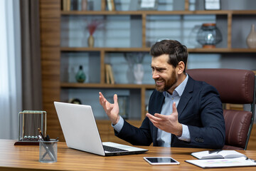 Obraz premium Angry and angry businessman talking on video call, talking to colleagues and shouting quarrel, mature boss working inside modern office building, senior man with laptop and beard.