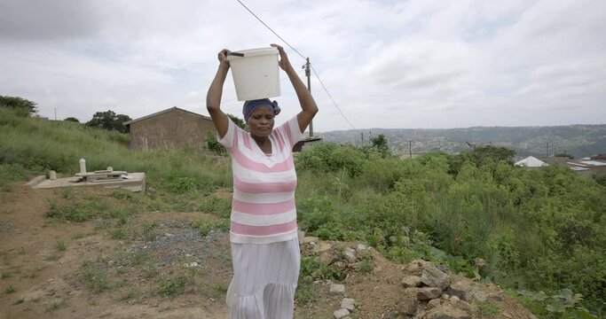 An elderly African woman carrying and balancing a bucket of water on her head in rural Zululand, South Africa.