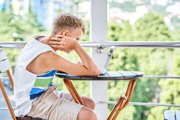 Preteen boy sitting on hotel balcony and surfing the internet on grey laptop. Schoolboy enjoys spending summer holidays and playing games