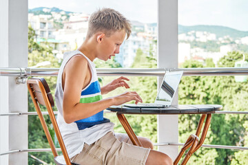 Preteen boy sitting on hotel balcony and surfing the internet on grey laptop. Schoolboy enjoys spending summer holidays and playing games