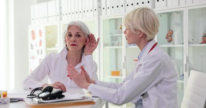 Mature woman patient shows sick ear to doctor sitting at table in clinic office. Medical professional checks client hearing ability slow motion