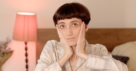 Joyful woman holds her face with her hands, poses, looks up. Graceful demeanor, relaxed manner. Against the background of a lamp in bokeh.