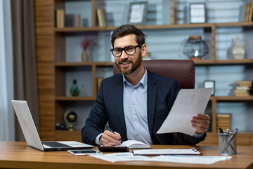 Obraz premium Portrait of mature businessman inside office at workplace, man with documents smiling and looking at camera, paperwork of mature experienced boss at desk.