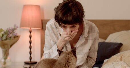 Allergic human reaction to cat hair. A woman sneezes while petting her pet in bed. Lamp light bokeh...