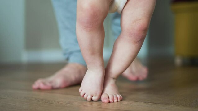 Charming child takes his first steps in his home, and happy parent is helping hand. Concept of happy family and home schooling.Develop your baby.First joyful steps of baby.Bare feet on parquet at home