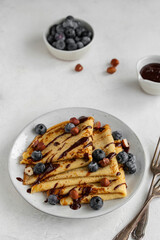Crepes, blinis or thin pancakes with blueberries, hazelnuts and chocolate sauce on a white plate at...