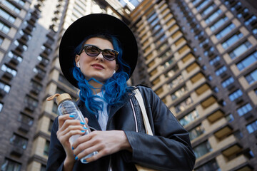 Portrait of a beautiful diverse person posing with glass water bottle by a high rise building in the city center. Stylish young female with dyed blue hair listening to music on a walk © hurricanehank