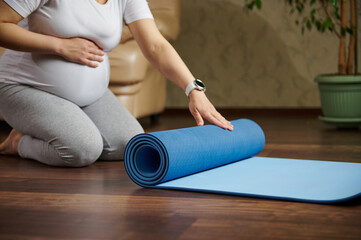 Selective focus on a blue exercise mat on a wooden floor, and a pregnant woman, gravid expectant mother rolling it out, for pregnancy yoga practice, breathing exercises for preparing to the childbirth