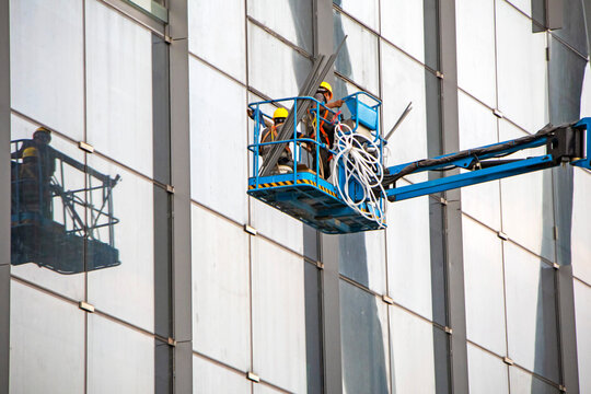 A construction worker in the bucket of a crane is installing insulation in the window block of the building. A man in the cradle, dismantling the old insulation of a sealed gasket to a new one.