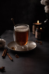 Freshly brewed black tea in a transparent glass with a steam on dark background with candles, cotton flowers and cinnamon sticks.