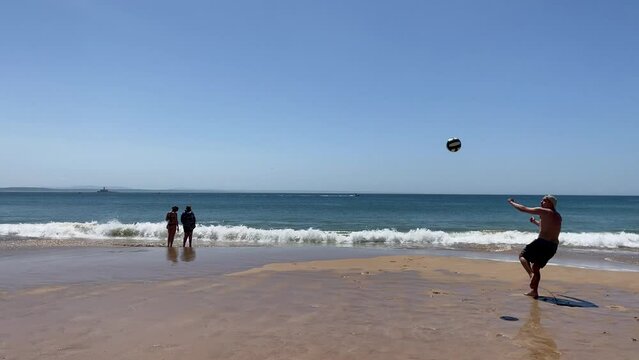 Two women wetting feet, bugio lighthouse in foreground, howler lamp and player kicking ball in background. Dynamic and playful scene enjoying a sunny day on cascais beach.