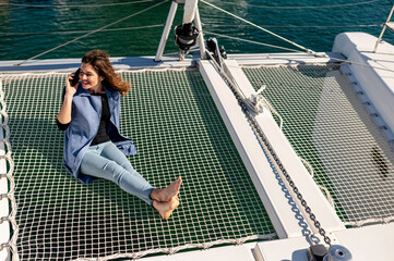 High angle of glad female sitting on net on yacht and talking on mobile phone during summer vacation