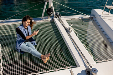 High angle of happy female sitting on net on yacht and browsing on mobile phone during summer vacation
