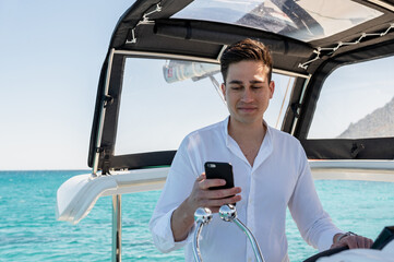 Cheerful male standing at steering wheel of catamaran and browsing social media on mobile phone...