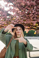 Pleased girl in black hat posing with flowers. Happy young caucasian girl with cute smile takes selfie against backdrop of flowering sakura tree in spring. High quality photo
