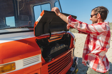 Side view of adult unshaven male traveler in casual clothes and sunglasses opening bonnet of retro van parked on sandy road during trip in nature