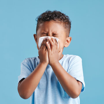 Sick, blowing nose and a boy with a tissue with covid isolated on a blue background in a studio. Health, young and a little boy with an allergy problem, virus and sneezing from a cold on a backdrop