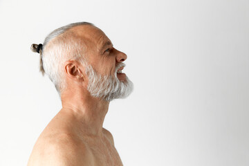 Profile view of handsome mature man with gray hair and beard posing shirtless, smiling against grey background. Stylish hairdo. Concept of male beauty, face and skin care, daily procedures, age