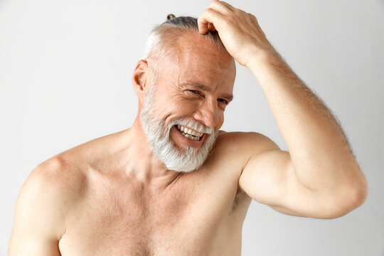 Portrait of handsome, mature, half-naked man with gray hair and beard smiling against grey background. Morning routine. Concept of male beauty, face and skin care, daily procedures, age
