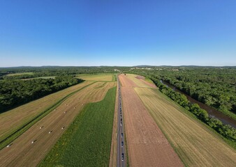 Aerial shot of a highway surrounded by fields, forest, and a river on a sunny day