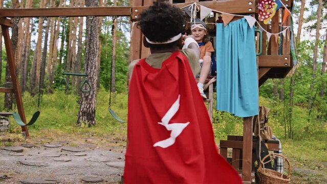 Medium shot of African American and Caucasian elementary age boys wearing costumes and roleplaying superheroes on playground next to treehouse