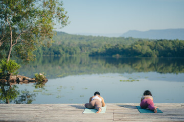 Fototapeta na wymiar Group of people doing yoga exercises by the lake at morning outdoor, Woman practicing yoga in lotus position at park, Multinational women doing breathing exercises or yoga meditation during outdoor