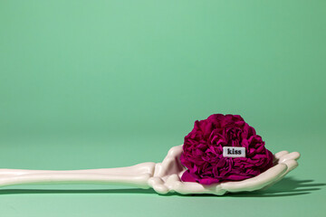 skeleton hand holding pink flower on green background, creative romantic concept, word kiss in flowers, art design

