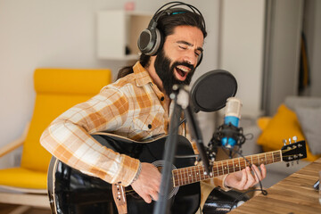 Fototapeta na wymiar Performing song. Inspired man, amateur music singer and writer, sitting in headphones, with guitar in at home sound recording studio. Young guitarist sings and plays live instrumental melody