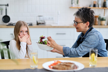 Little girl child closes her mouth and does not want to eat vegetable salad for lunch, adult mother...