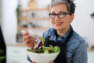 Portrait of a smiling mature woman in glasses with a large plate of salad in the kitchen.
