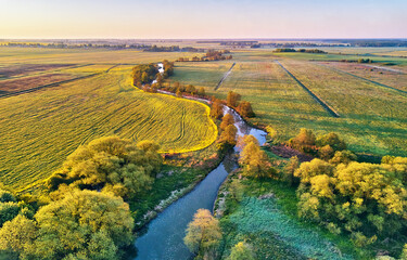 Spring morning landscape. Calm river in blooming meadows. Aerial rural view from above. Agricultural yellow green colza fields. Rapeseed and dandelion blossom scene - 606001299