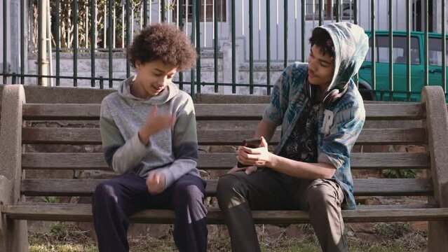 Two mixed race african teen boys sitting on park bench. The older boy shows the other something on his phone, which causes him to cringe with embarrassment.
4K 25fps
