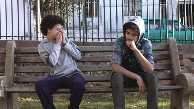 Two mixed race african teen boys sitting on park bench. The one boy tells the other one that he smells and moves away. The remaining boy smells his armpits to see what the problem is.