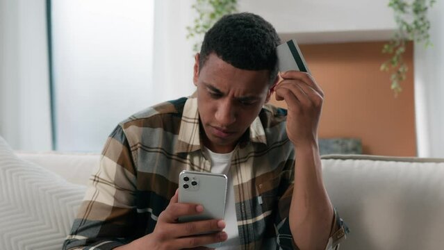 African American man frustrated confused guy at home couch problem with online payment with phone and credit card bank failure try to pay e-bank app error no money scam fraud unsuccessful transaction