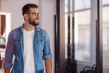 Cheerful entrepreneur in denim shirt, wearing glasses, standing in his office in the morning.
