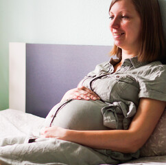 Happy smiling pregnant woman sitting on a bed in a bedroom at home and hugging her belly.