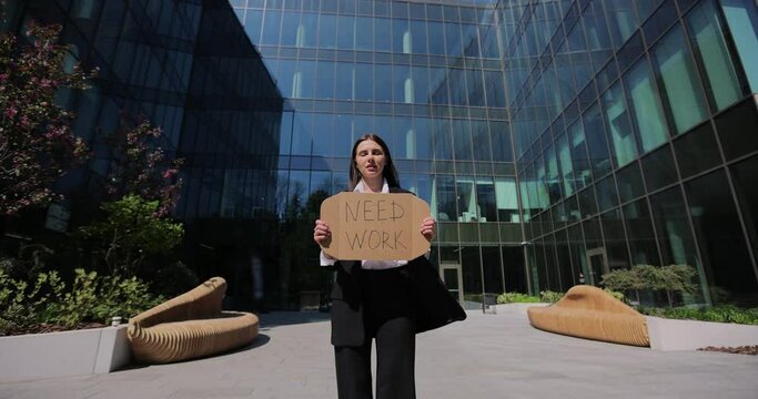 Businesswoman in a black suit holding Need Work sign searching job standing near office centre. Jobless people concept. Global unemployment.