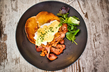 Top view of egg Benedict served with fried bread, bacon, and lettuce on a black plate